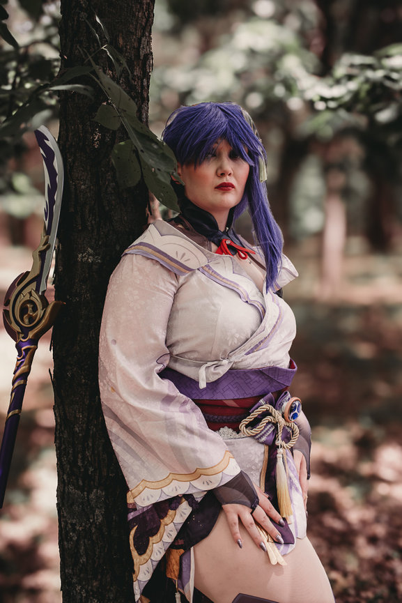 Person in purple anime-style cosplay, leaning against a tree in a forest. Cosplay includes a detailed sword and kimono.