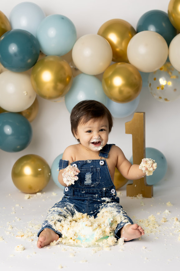 Baby wearing denim overalls, sitting in cake with a joyful expression, surrounded by balloons and a 1 sign.