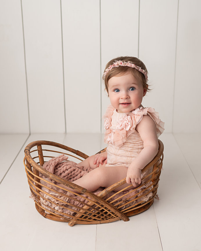 One-year-old sitting on natural wood basket in minimalist white studio