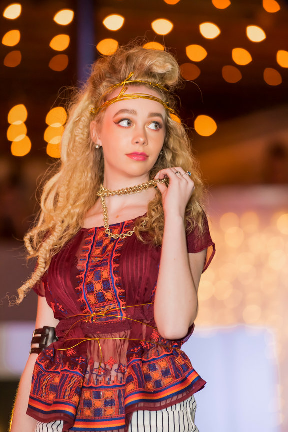 Fashion model on runway wearing a burgundy blouse with intricate orange and blue embroidery, paired with striped pants, gold chain necklace, and gold wire headpiece, against a background of warm bokeh lights.