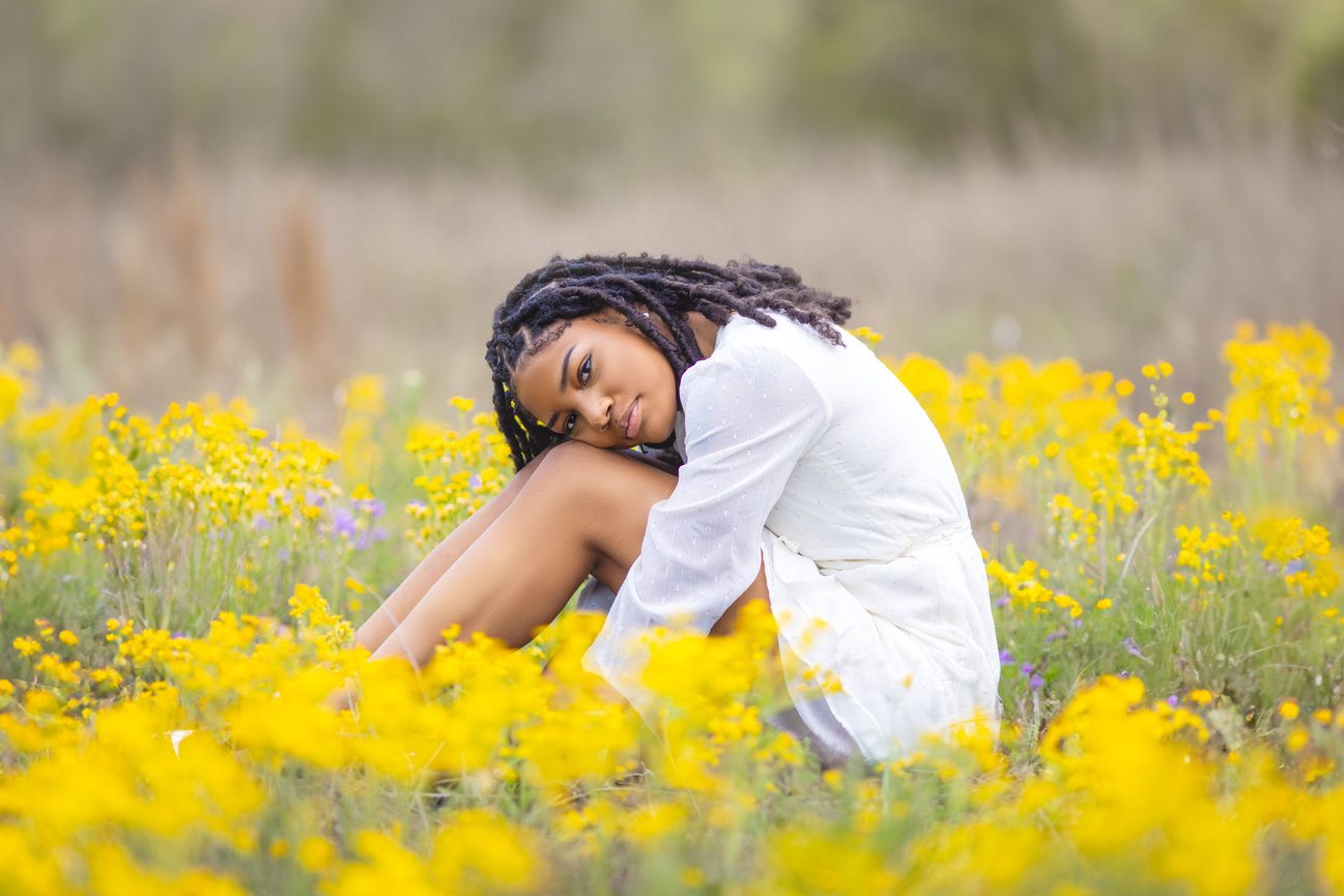 Graduate sitting in a field of yellow flowers, dressed in a white dress at Erwin Park, Texas.