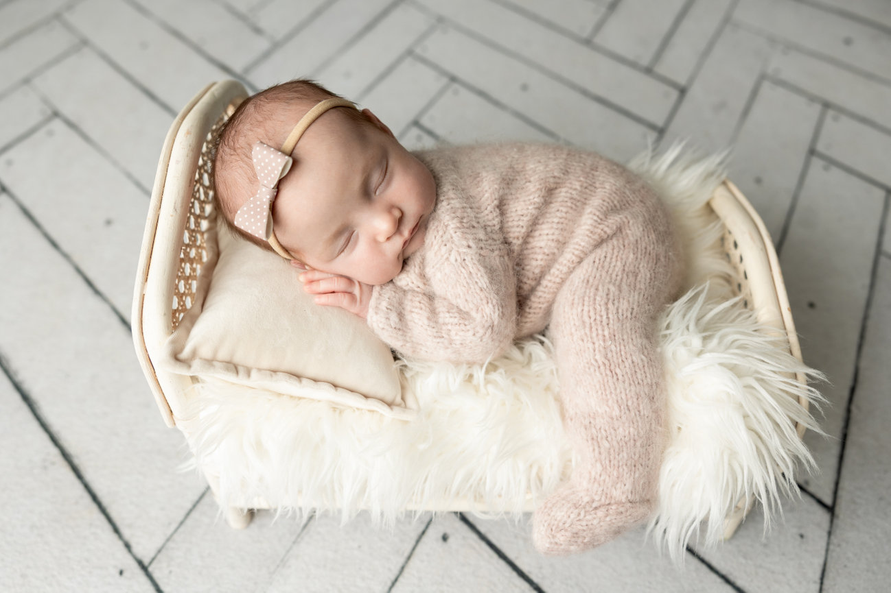 Sleeping baby in a knitted outfit, resting on a small bed with fluffy bedding.