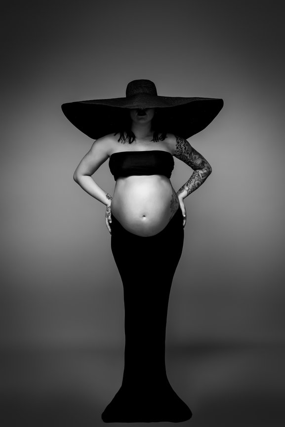 Pregnant woman in a black dress and large hat, posing with hands on hips against a neutral background.