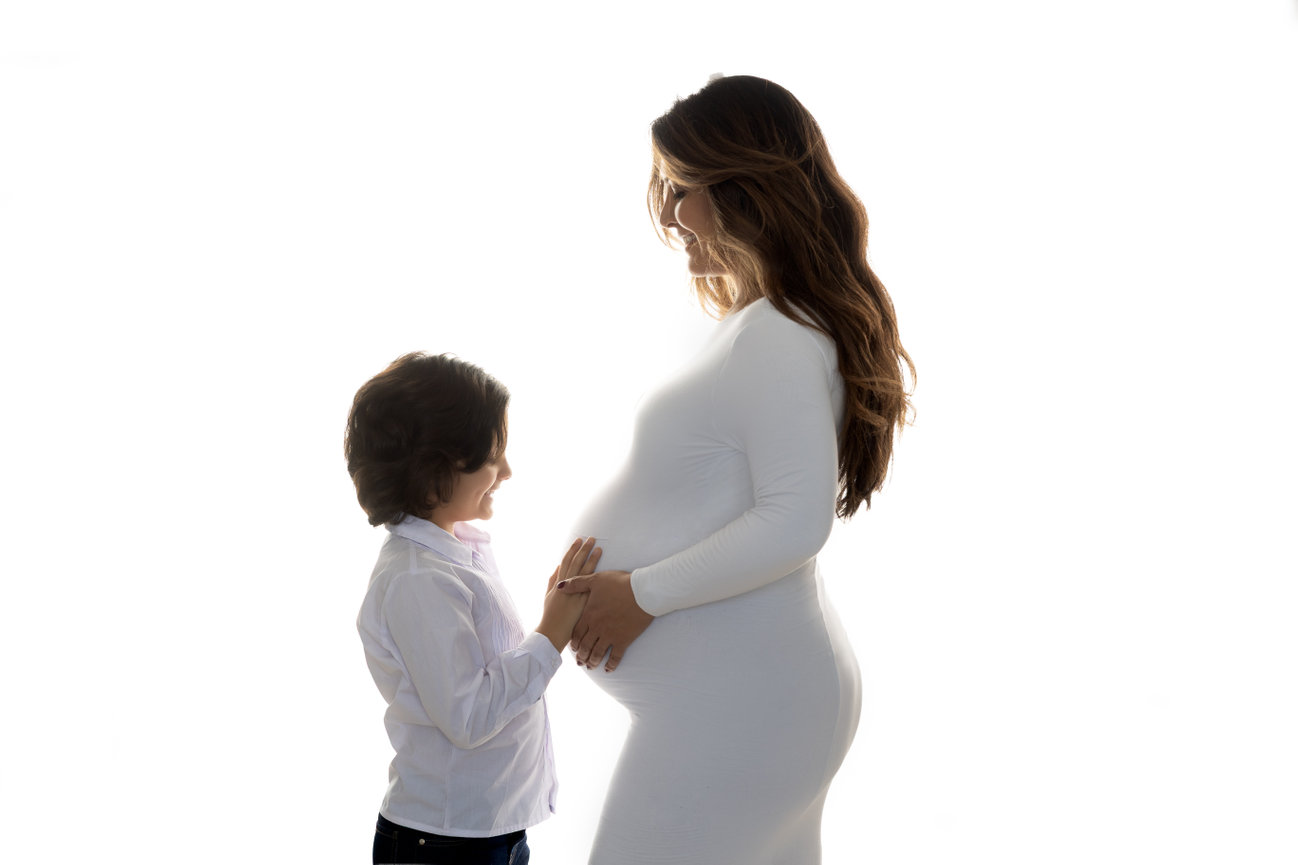 Pregnant woman in white dress smiling, with a child touching her belly, against a white background.