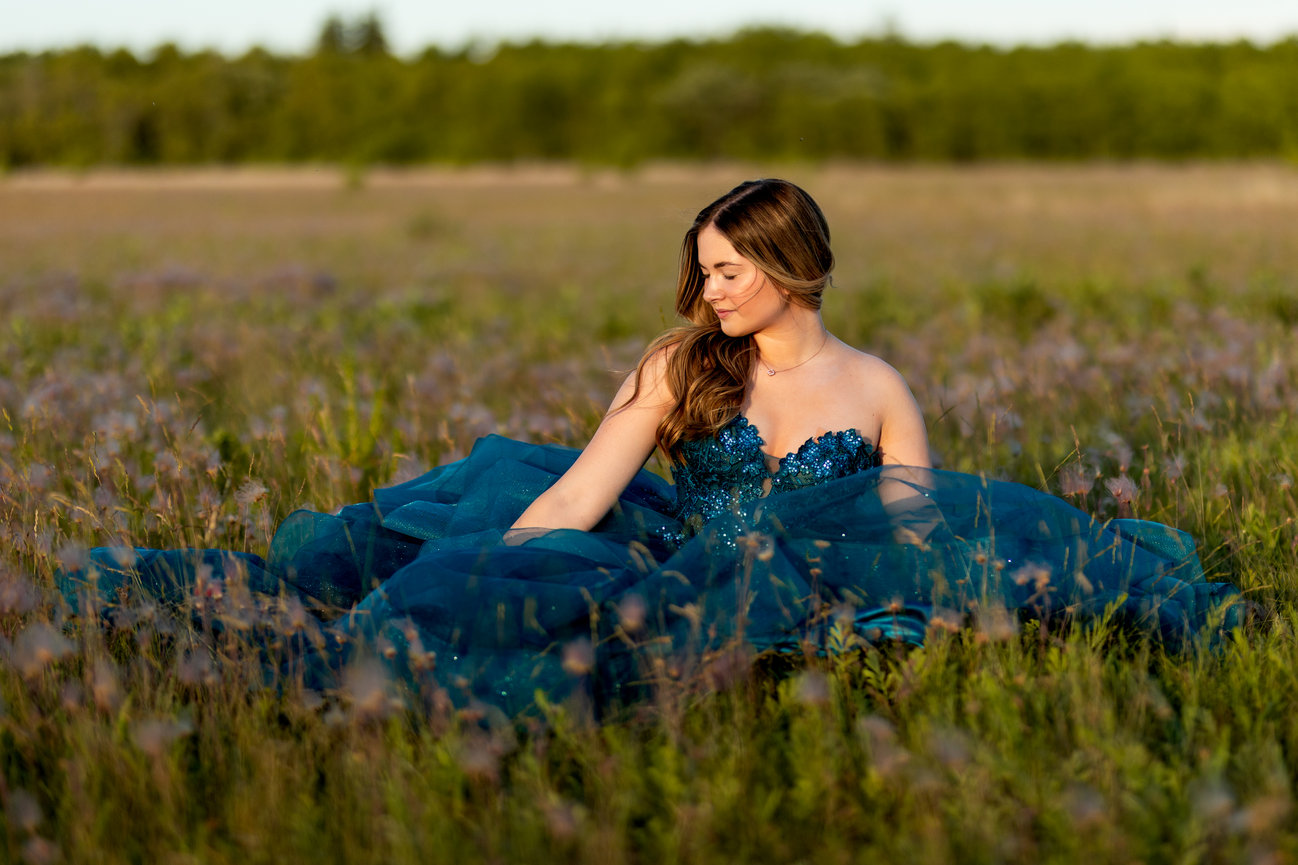 A woman in a blue gown sits in a grassy field at sunset, surrounded by wildflowers.