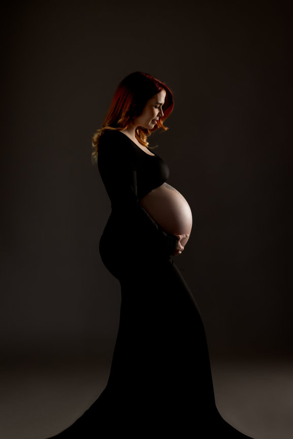 Pregnant woman in a long black dress poses against a dark background holding her belly.