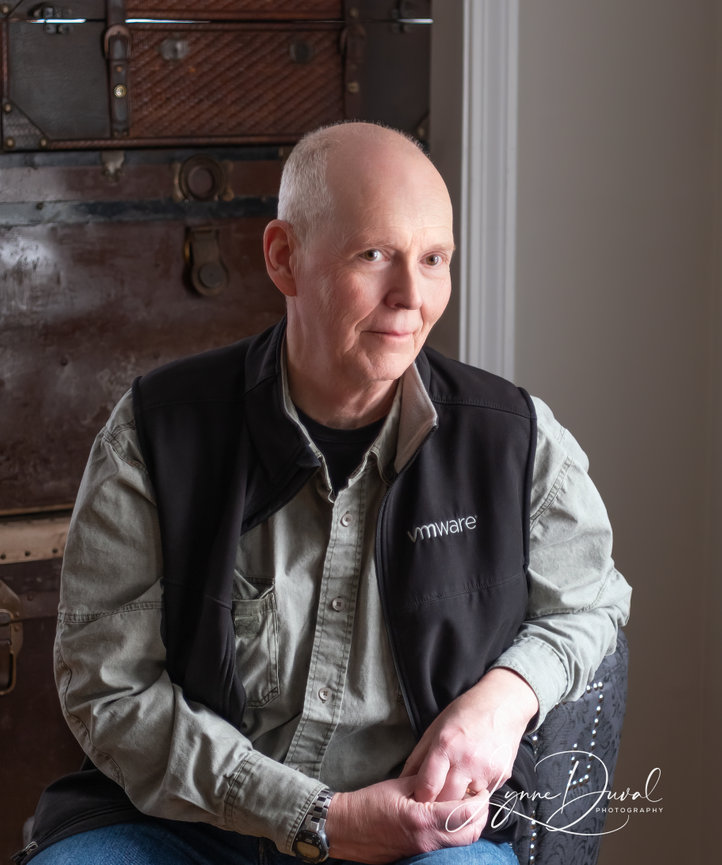 Bald man sitting down wearing jeans, black vest, and green long sleeve shirt