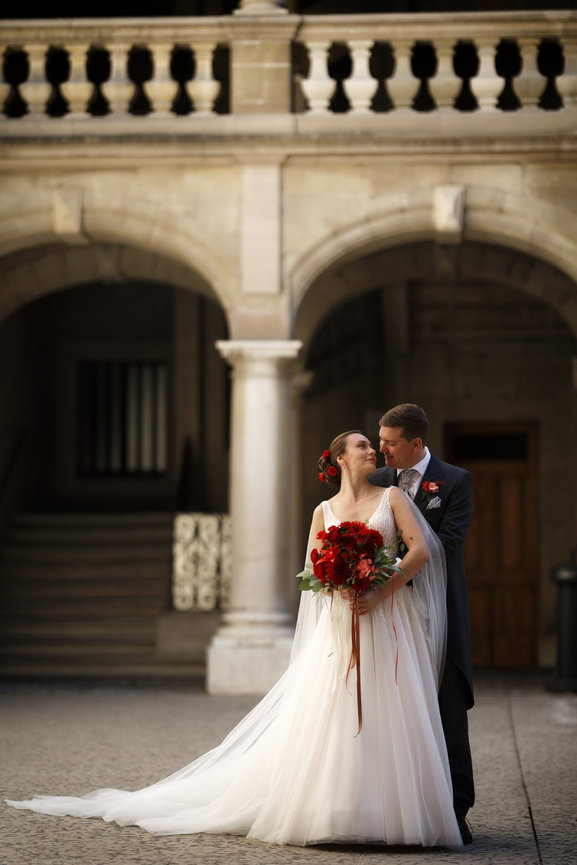 Bride and groom embracing in a stone courtyard beneath an arched balcony with a flowing white gown and red bouquet