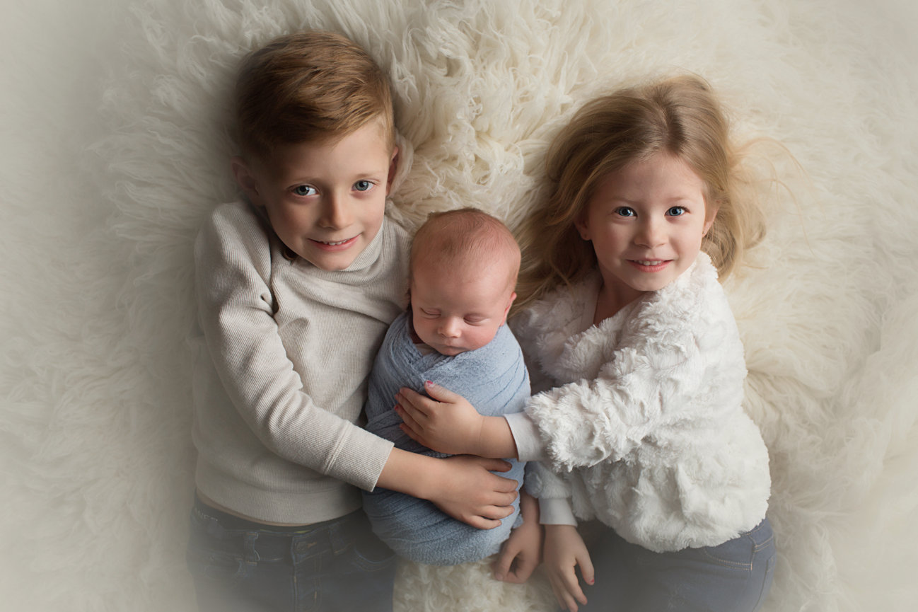 Three siblings laying on white fur with newborn sibling sleeping safely in the middle during newborn photoshoot with Kansas City newborn photographer Dreamworks Photography 
