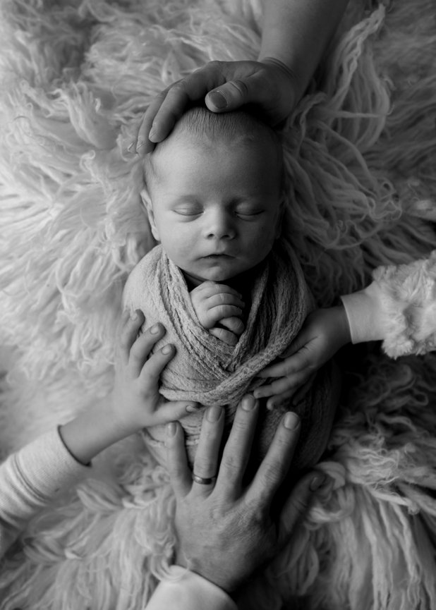 Black and white image of newborn baby sleeping with one hand of each family member placed on him including mom, dad, and two siblings with Kansas City newborn photographer Dreamworks Photography