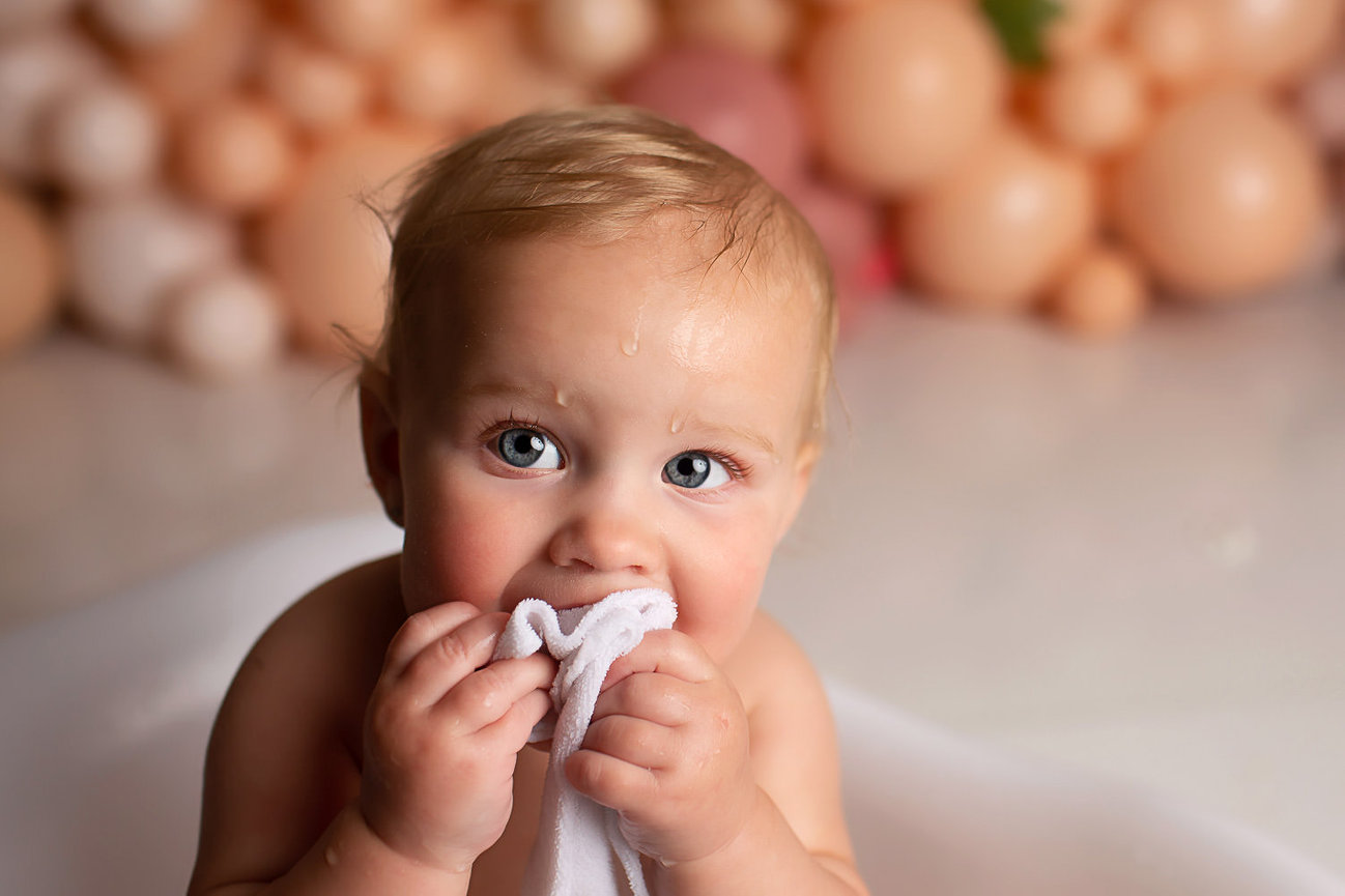 Closeup of baby girl with white washcloth in her mouth in a tiny baby tub in front of peach colored balloon backdrop during first birthday cake smash photography session with baby photographer Dreamworks Photography in Kansas City photography studio