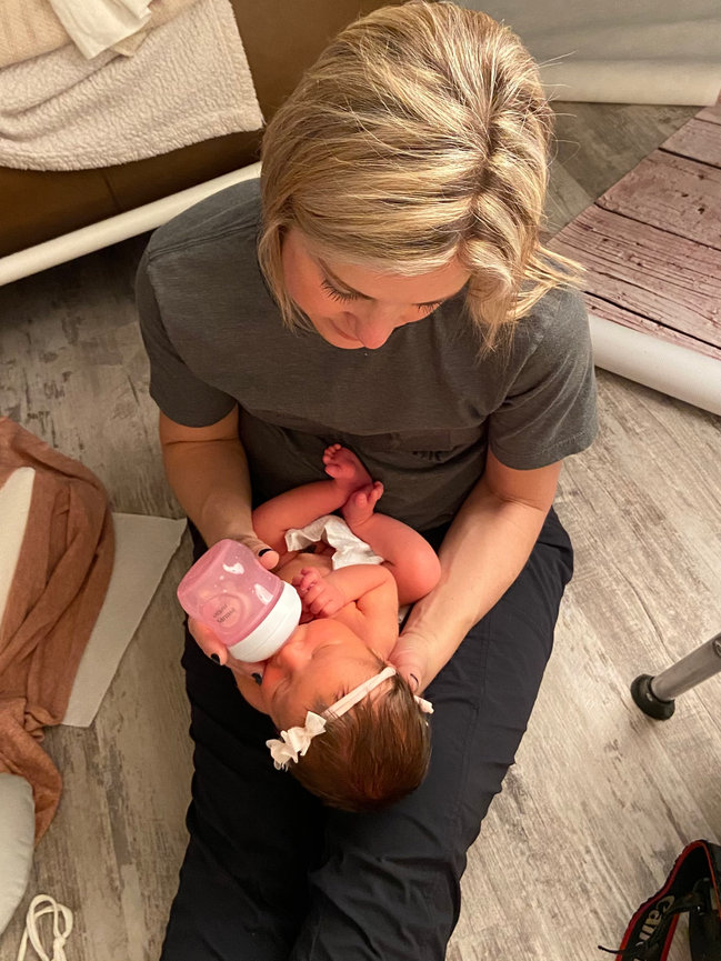 Omaha newborn photographer sitting on the floor feeding a baby with a bottle, surrounded by soft blankets.
