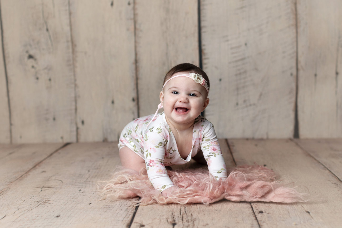 Baby in floral outfit and headband smiling on wooden floor with pink fur rug.