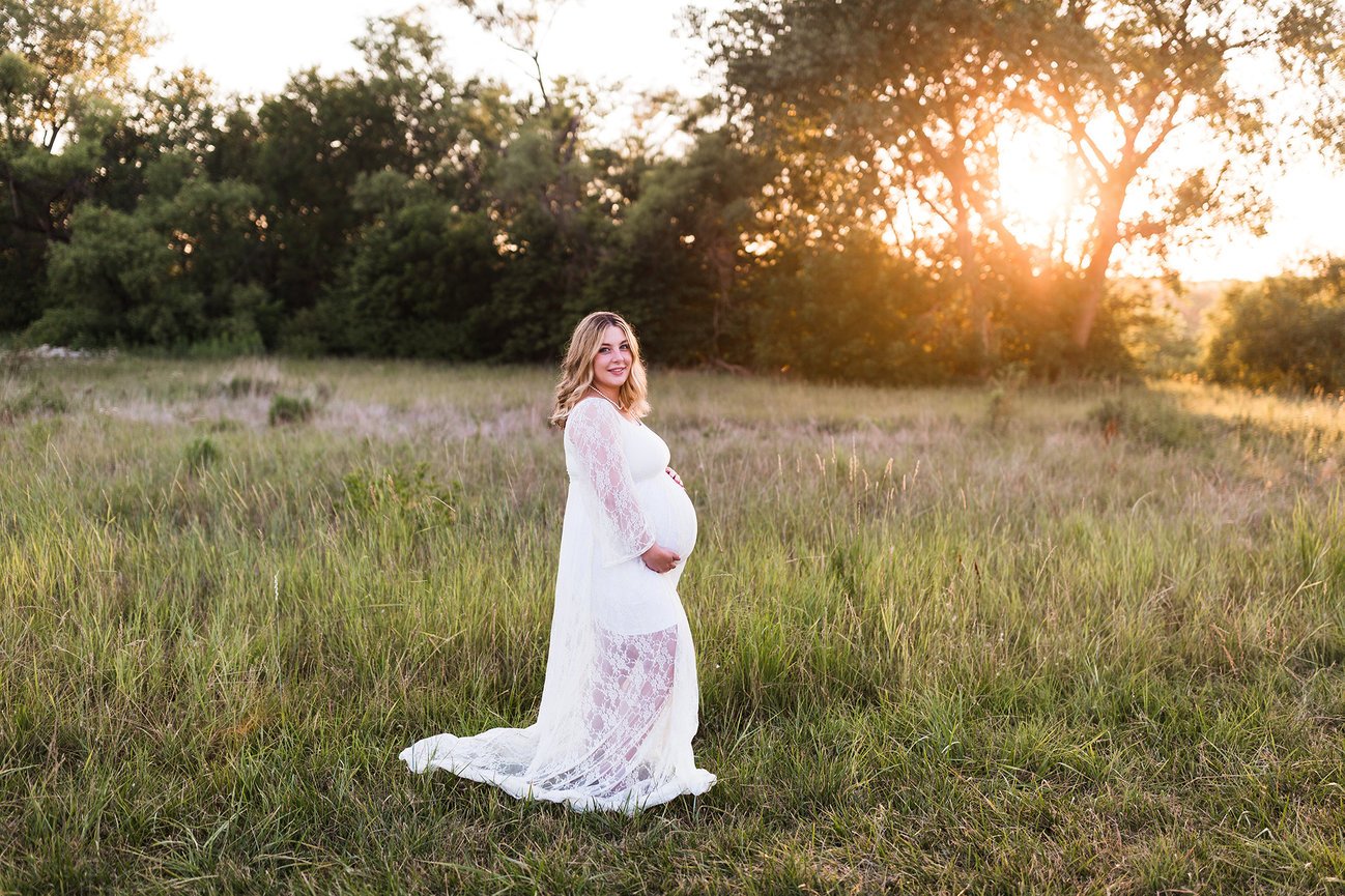 Pregnant woman in a white lace dress stands in a grassy field at sunset.