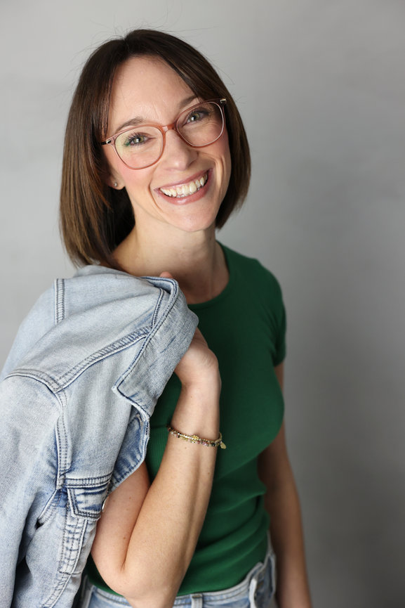 Smiling woman in glasses holding a denim jacket over her shoulder against a gray background. Business content photos melb