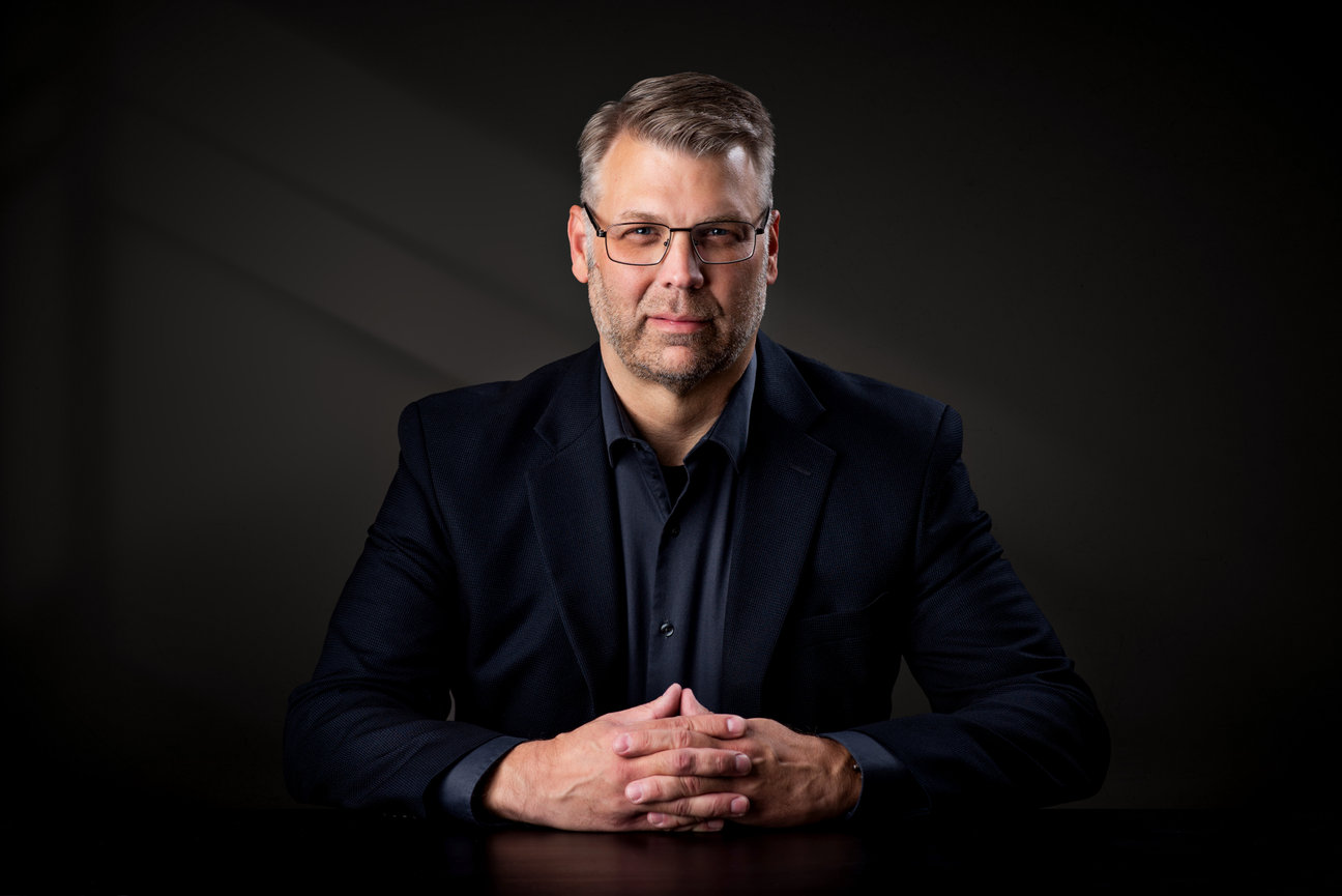 Man in a suit sitting at a table in a dark studio posing for a headshot.