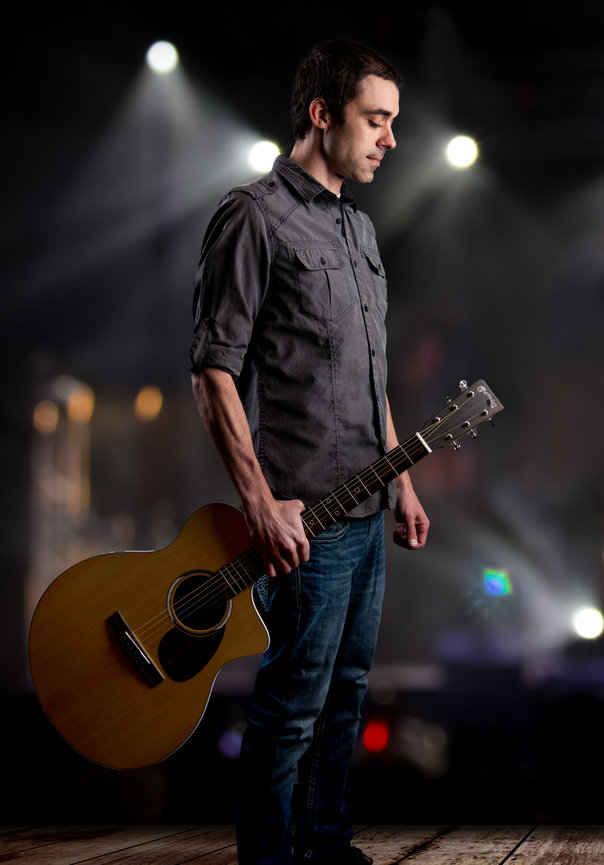 Man holding a guitar on stage looking down posing for a commercial photo shoot.