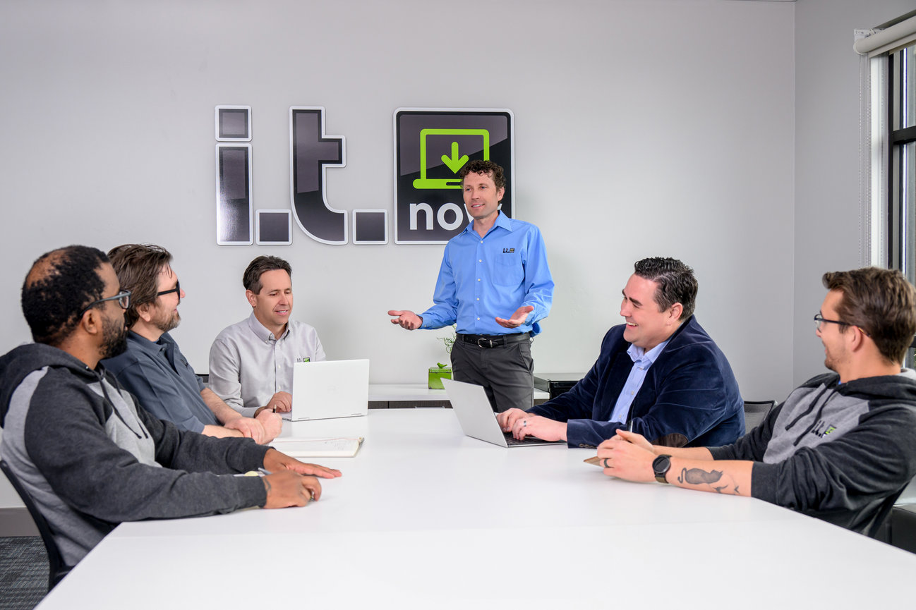 A team of six professionals in a meeting room discussing IT solutions with laptops and a presentation.