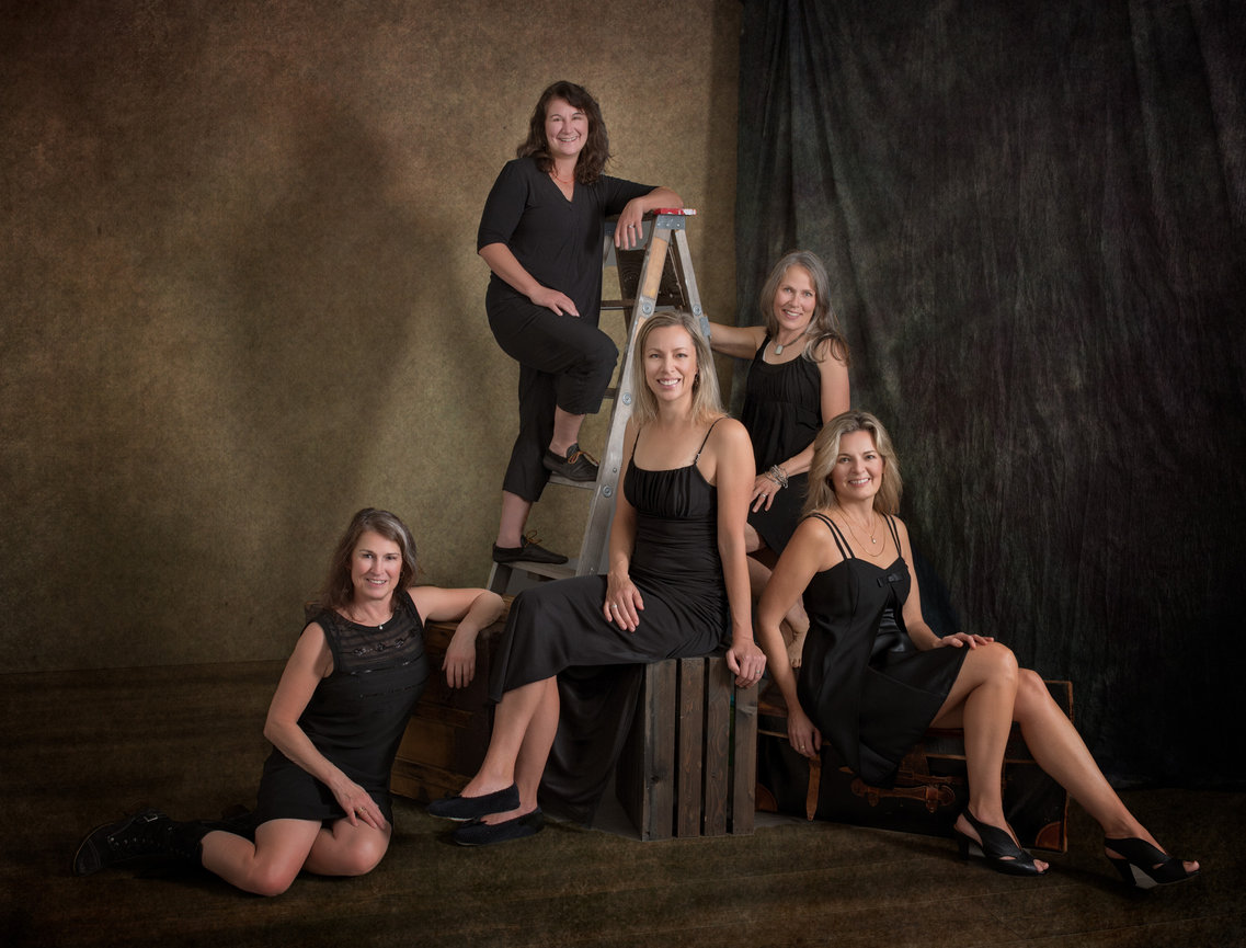 Five women in black dresses pose in a studio portrait, seated and standing around a ladder and crates.