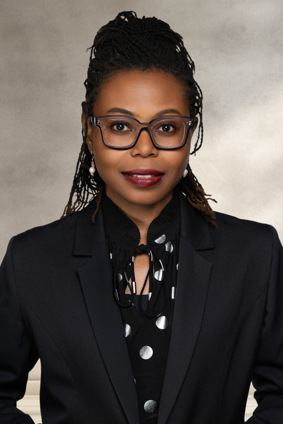 Professional portrait of a woman with glasses, wearing a stylish black suit and a polka dot blouse, against a neutral background.