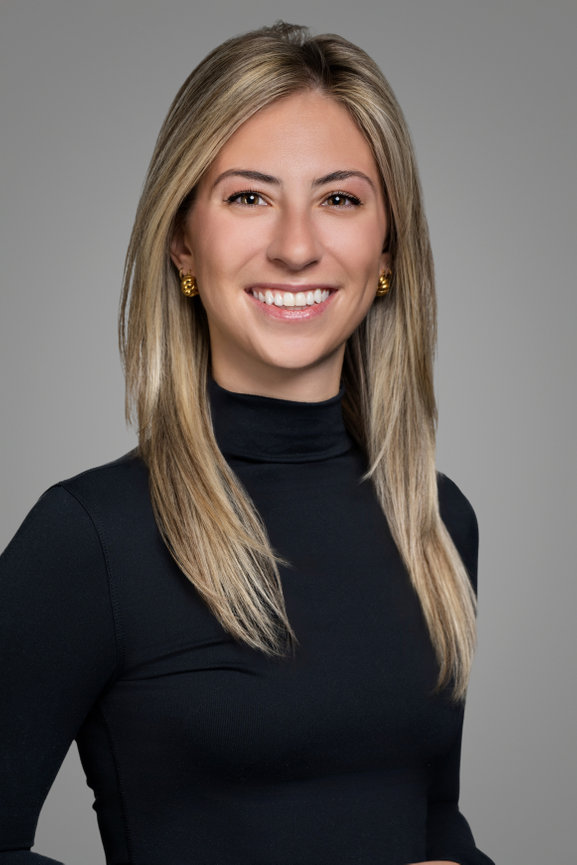 Woman with long blonde hair, smiling, wearing a black turtleneck and gold earrings against a gray background.