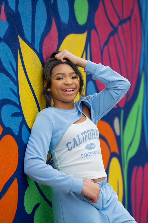 Smiling person in a light blue outfit leans against a colorful mural with floral patterns captured by Jo Hayes Images Charlotte Portrait Fashion Photographer