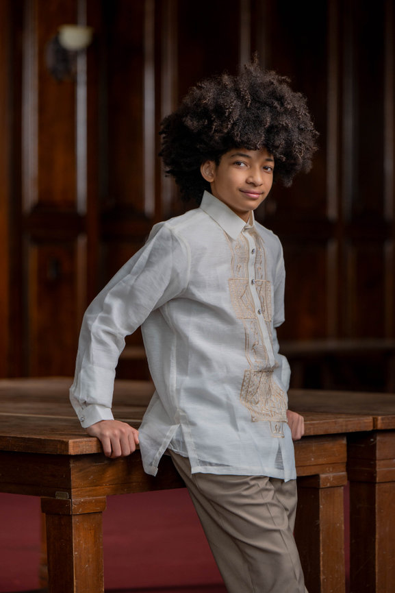 Person with curly hair in a white embroidered shirt and beige pants, typical filipino barong, leaning against a wooden table, with a wood-paneled background in editorial style captured by jo hayes images charlotte portrait photography