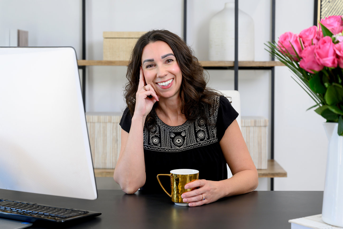 Smiling person at a desk with a computer and coffee, beside a vase of pink flowers.