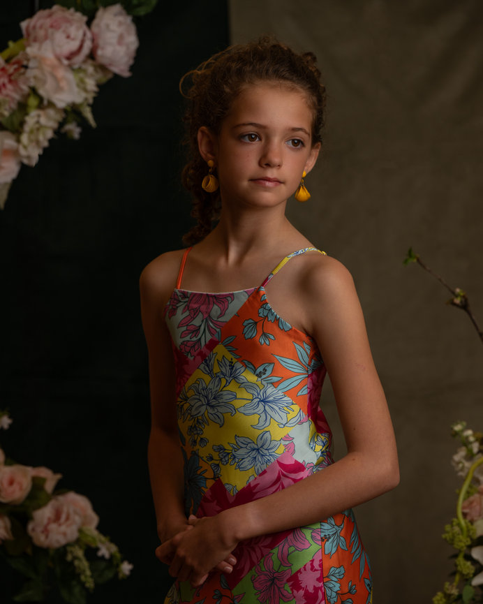 Young girl in floral dress with curly hair and yellow earrings, standing among pink flowers, gazing thoughtfully captured by Jo Hayes Images Charlotte Portrait Photography