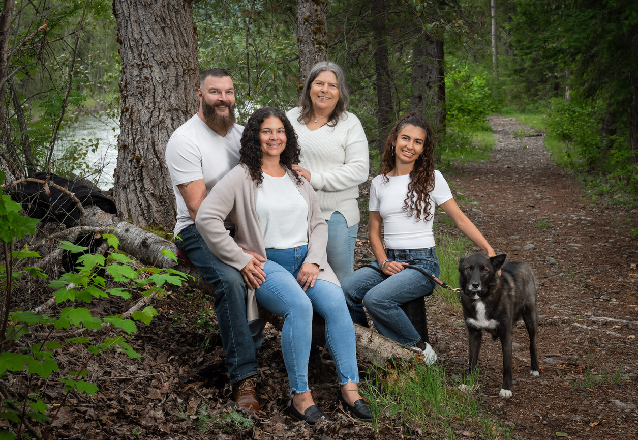 Family portrait with four people and a dog in a wooded area beside a river, wearing jeans and white shirts.