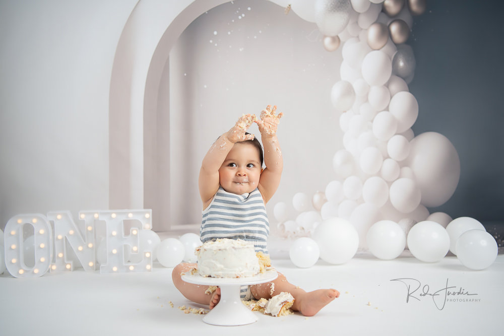 A smiling baby in a striped outfit holds up a crown next to a white cake and festive balloon decorations.