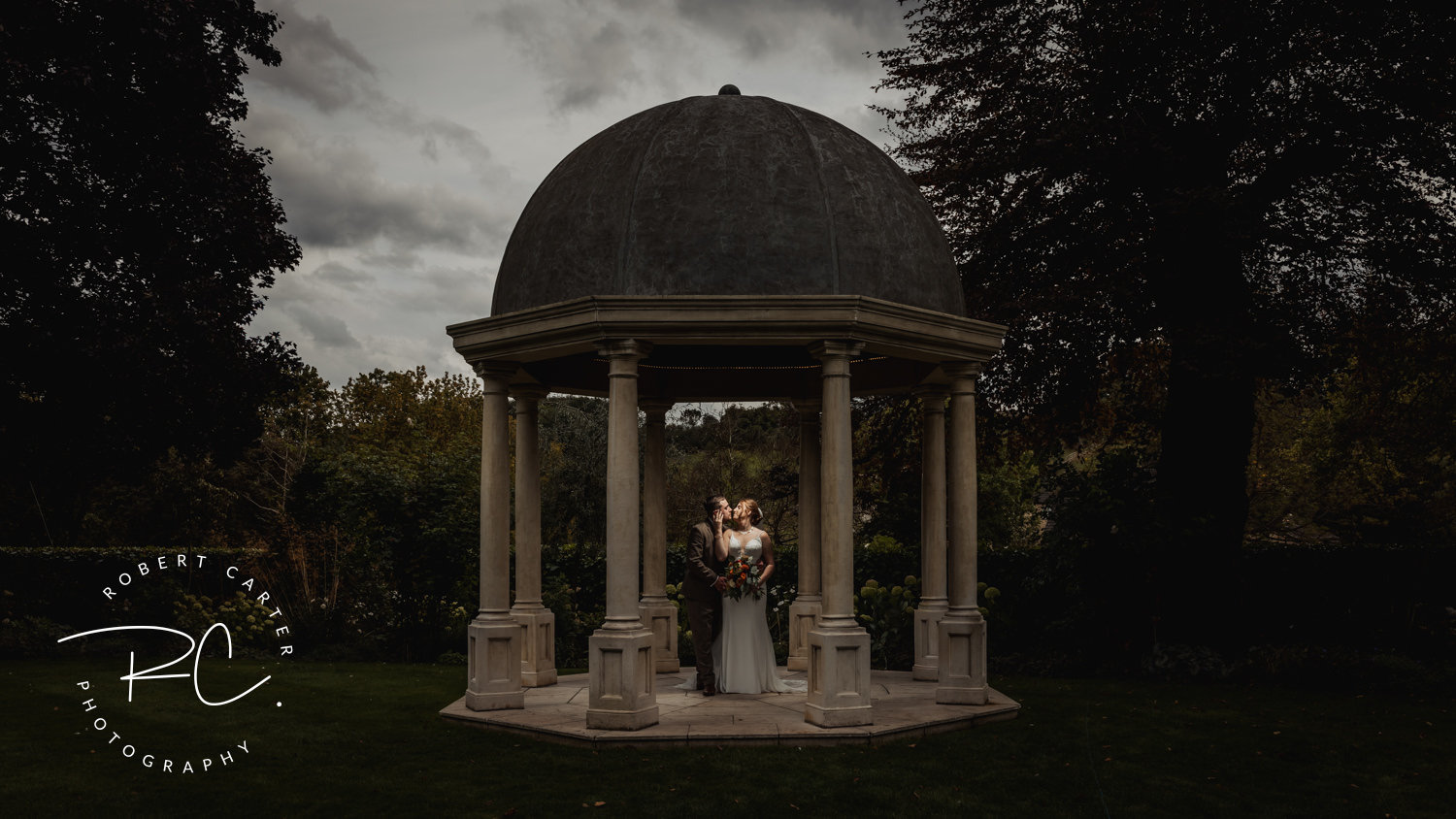 Bride and groom stand inside a gazebo, surrounded by trees, under a cloudy sky.