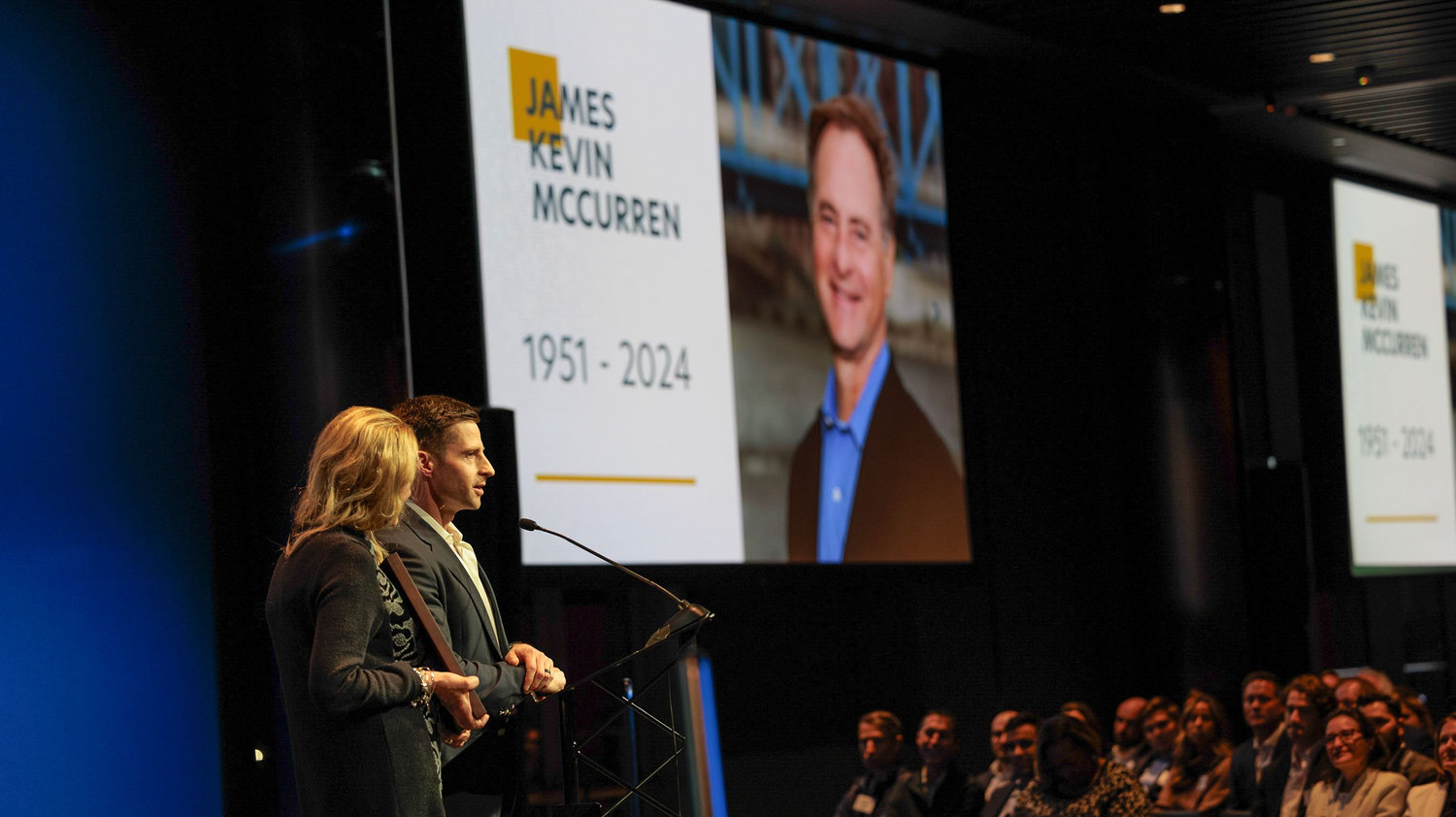 A man and woman speak at a podium next to a large screen displaying an image and the name James Kevin McCurren 1951-2024 at a memorial event.