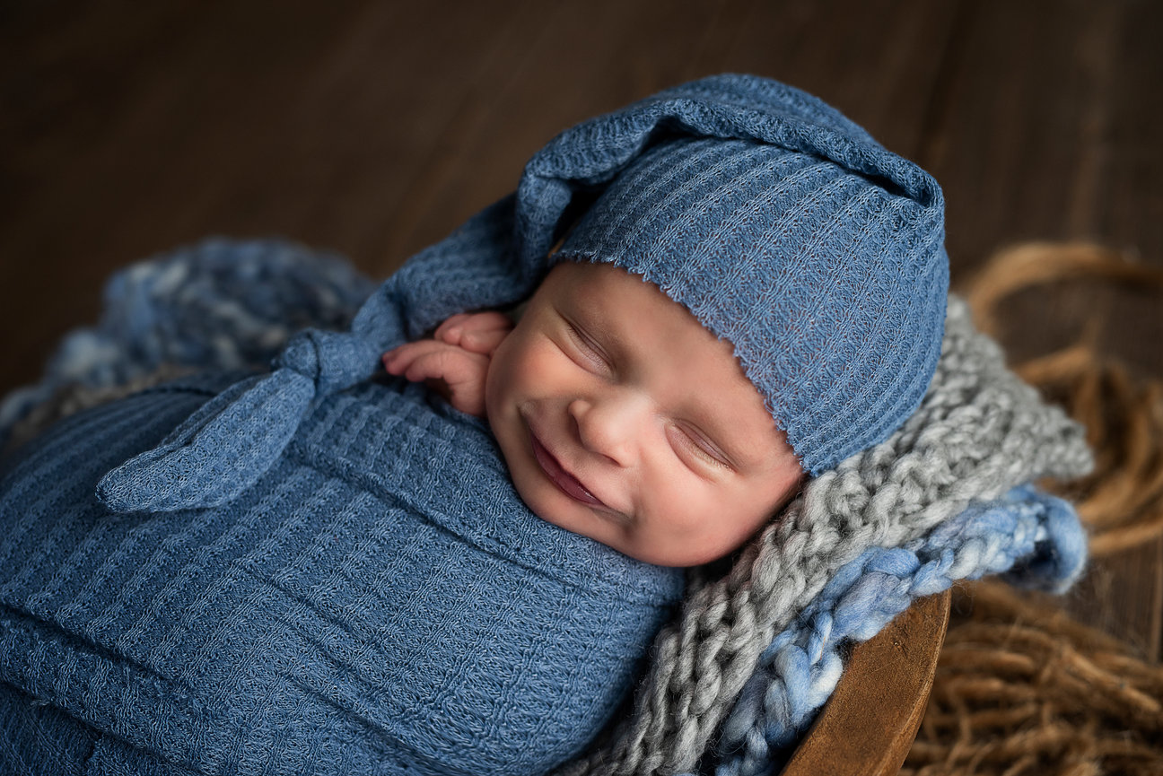 A newborn baby is peacefully sleeping, wrapped in a cozy blue knitted blanket with a matching hat. Nestled in Schaumburg, Illinois, the baby smiles softly on a textured grey and brown surface, creating a warm and serene atmosphere.