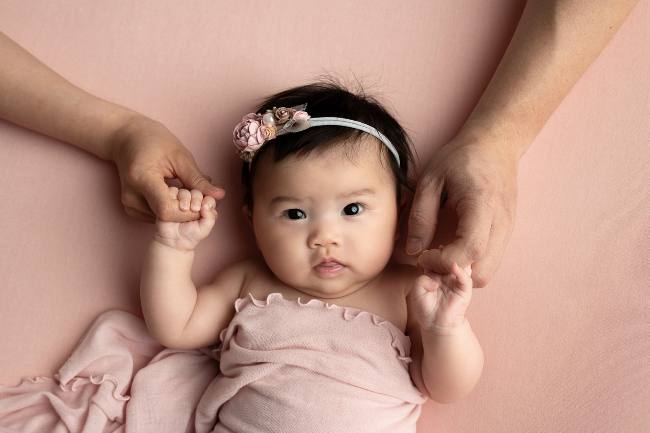 A baby wrapped in a soft pink blanket lies on a matching background. The baby wears a floral headband and looks directly at the camera. Two adult hands, as tender as the spirit of Chicagoland, gently hold the babys hands.