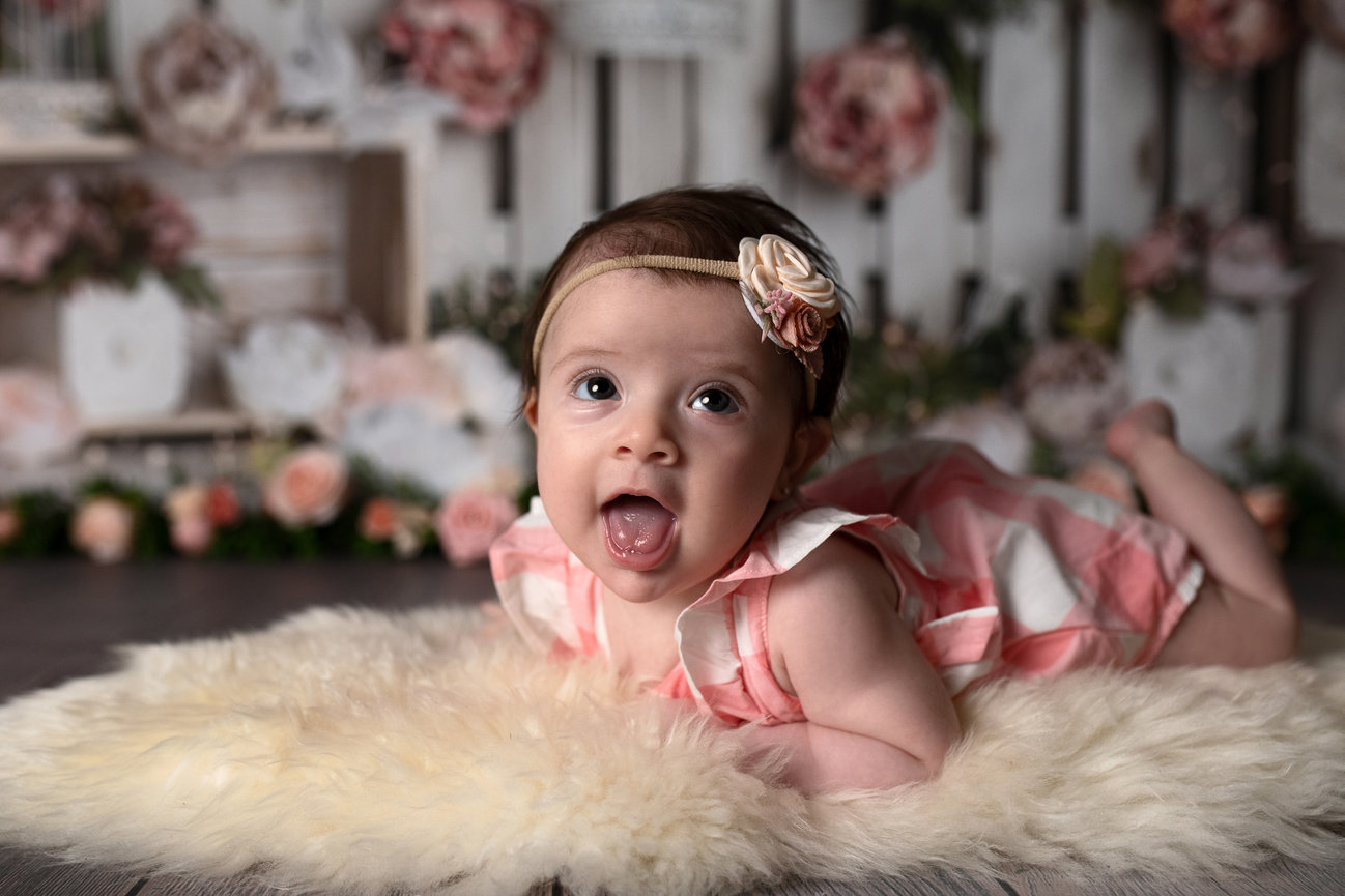 A baby is adorably nestled on a soft, fuzzy rug, wearing a pink and white dress with a floral headband. Embracing the essence of child photography, the background features floral decorations in soft pink and white tones, creating a cozy atmosphere.