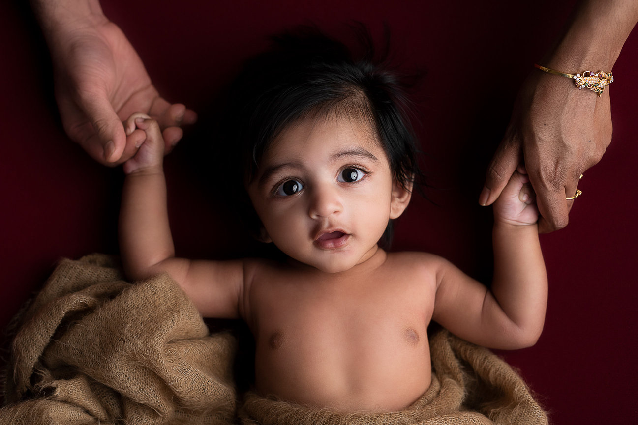 A 3-month photography session captures a baby with dark hair lying on a red background, wrapped in a brown blanket. The baby clutches an adults hand on each side, the right displaying a gold bracelet.