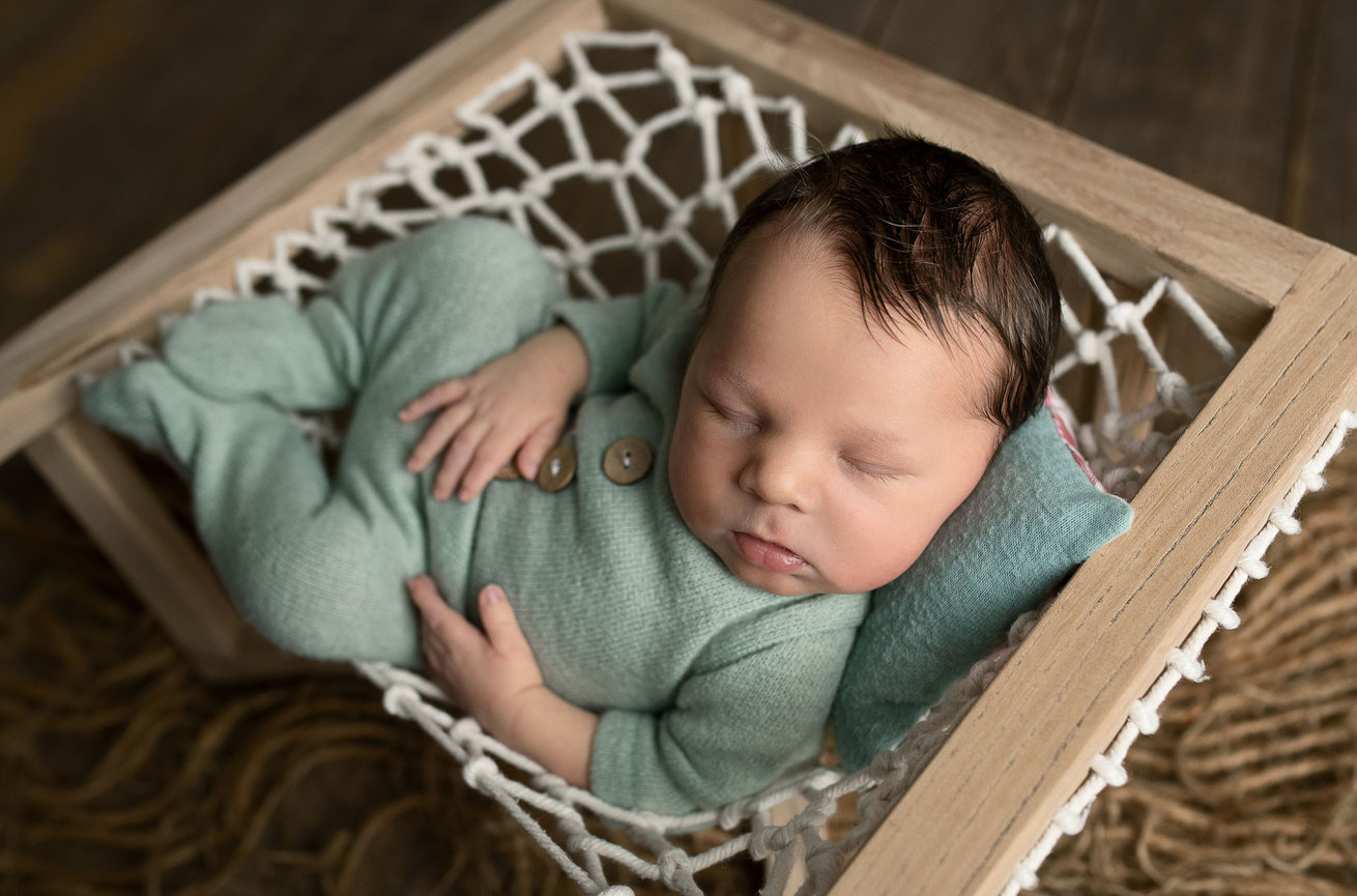 In Burlington, a newborn baby sleeps peacefully in a wooden-framed hammock. Dressed in a light green outfit with buttons, the baby rests on a small green pillow. The scene is set against wooden flooring with a brown woven mat beneath the hammock.