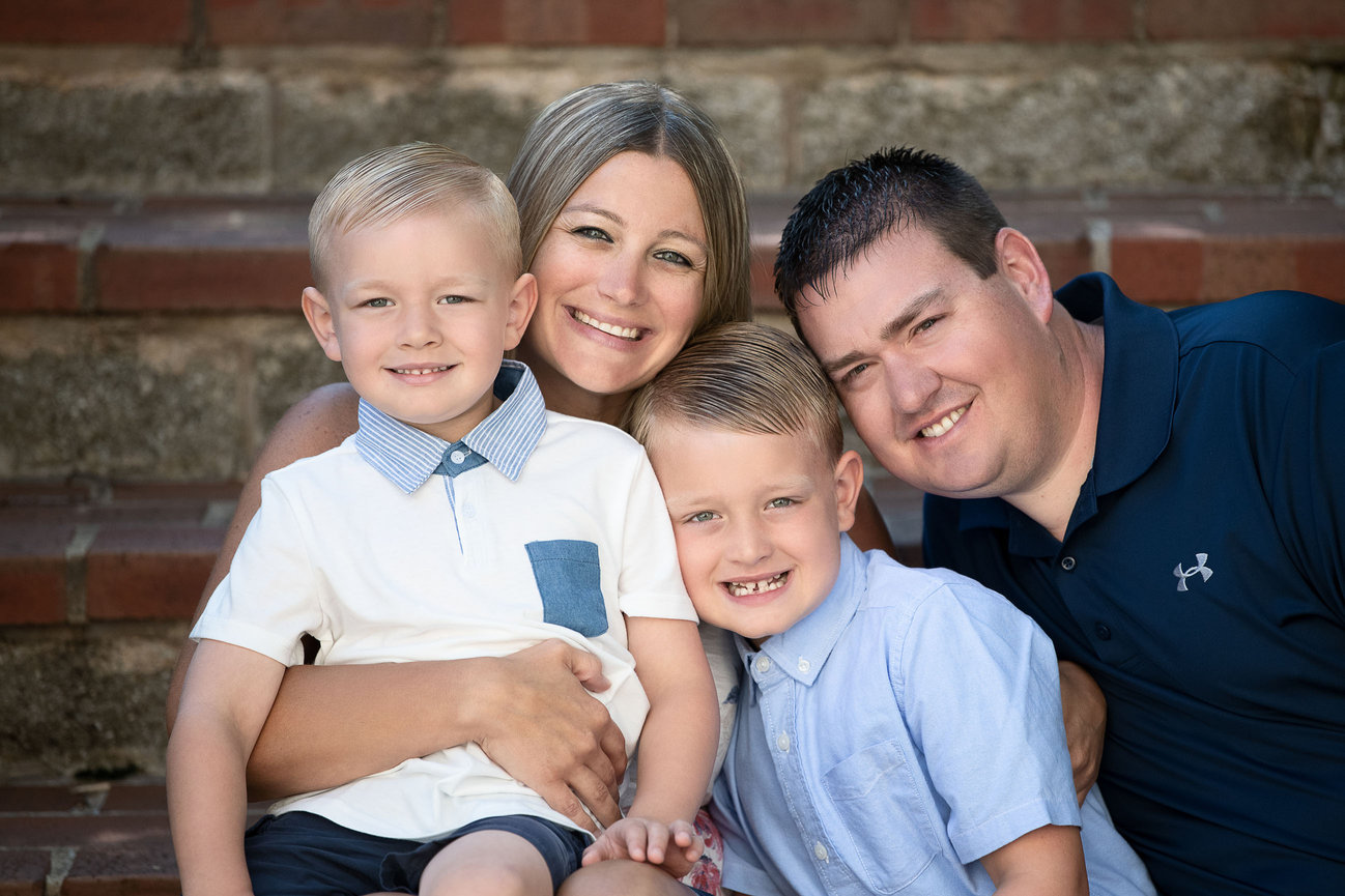 Captured by a Streamwood photographer, a family of four sits smiling on brick steps. The mother with long hair and the father are behind their young boys, who sport collared shirts and short hair.