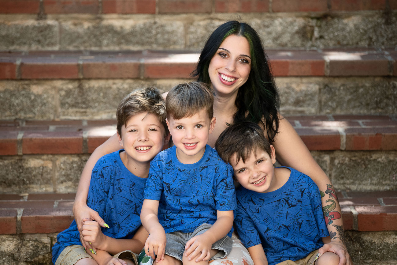 A woman sits on brick steps in Burlington, Illinois, with three young boys wearing matching blue shirts. They are all smiling at the camera, capturing a perfect moment of joy through photography, with their arms wrapped around each other.