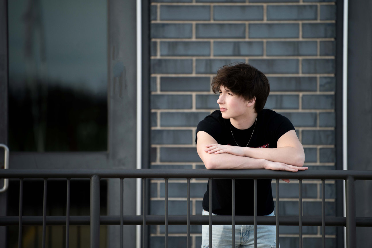 A senior with short brown hair leans on a railing, looking to the right. They sport a black T-shirt and light blue jeans. A brick wall and window form the backdrop.