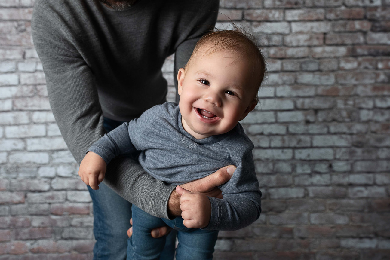 A smiling baby in a gray shirt is joyfully held up by an adult wearing similar attire during a 1st birthday photo. The rustic white brick wall in the background complements the playful mood, capturing a moment of pure happiness.