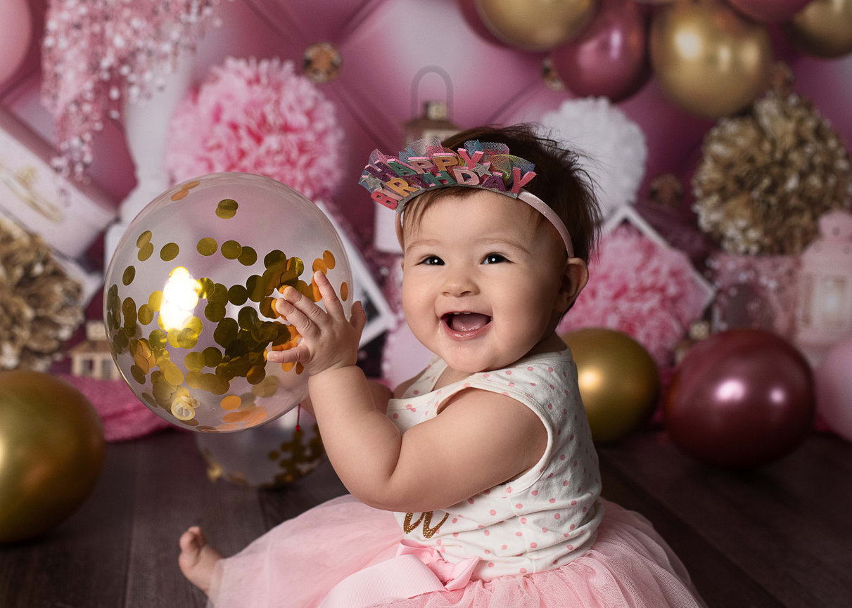 A baby wearing a Happy Birthday headband is sitting and smiling, holding a clear balloon with confetti. The background features pink and gold decorations.