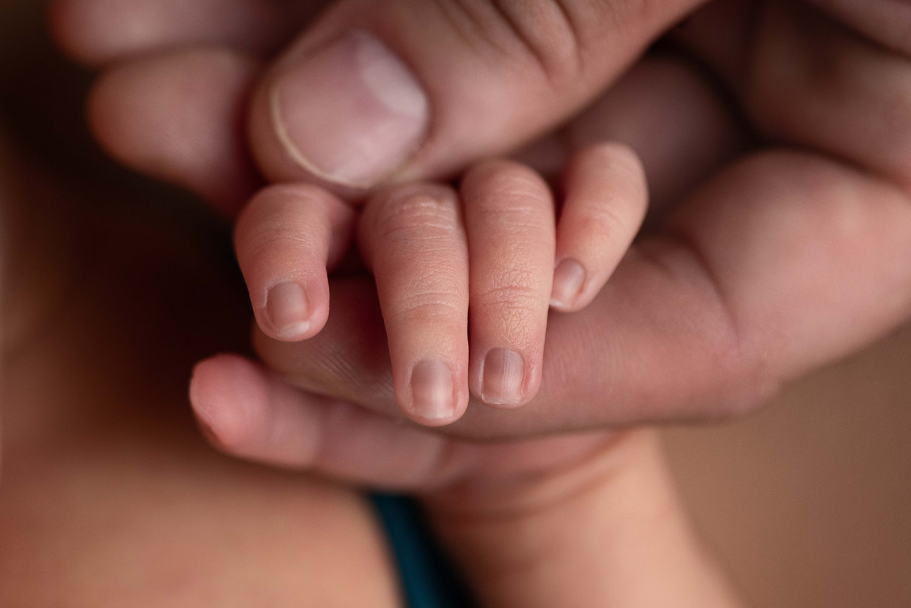 A close-up image shows an adult hand gently cradling a babys tiny hand, highlighting the small, delicate fingers of the baby compared to the adults. The softly blurred background beautifully emphasizes this tender connection between parent and child.