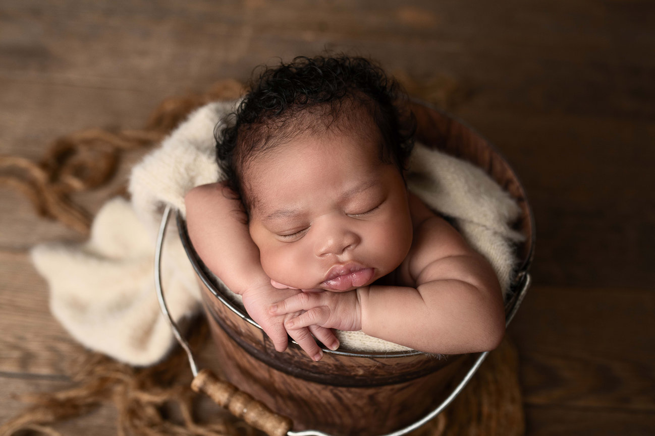 A recién nacido peacefully sleeps in a wooden bucket, with tiny arms resting on the rim. A soft blanket is draped inside, and the background features a wooden floor with a charming rope accent.