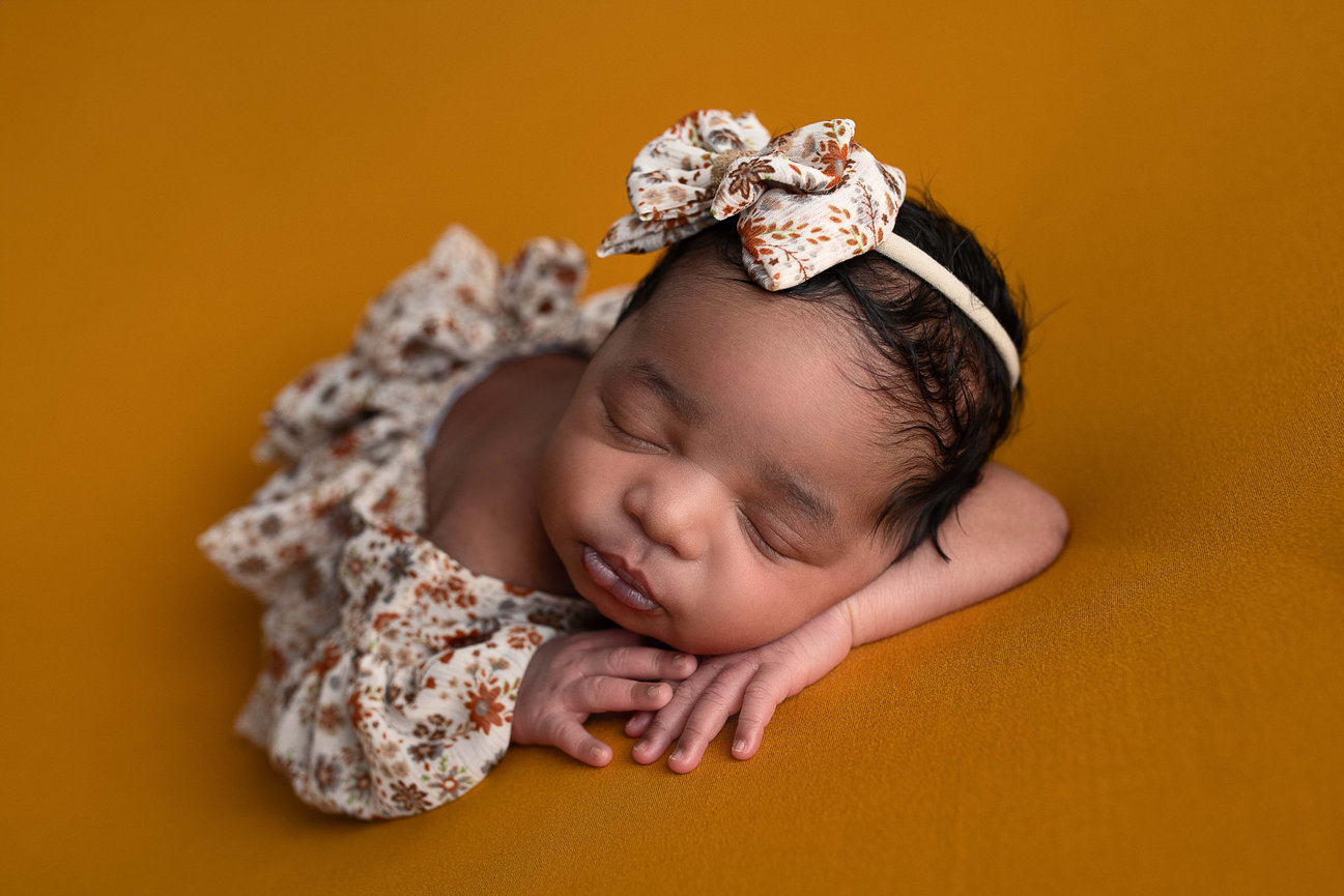 A newborn baby sleeps peacefully on a mustard yellow blanket in Marengo, Illinois. The baby wears a floral outfit with a matching bow headband and rests with their hands tucked under their chin.