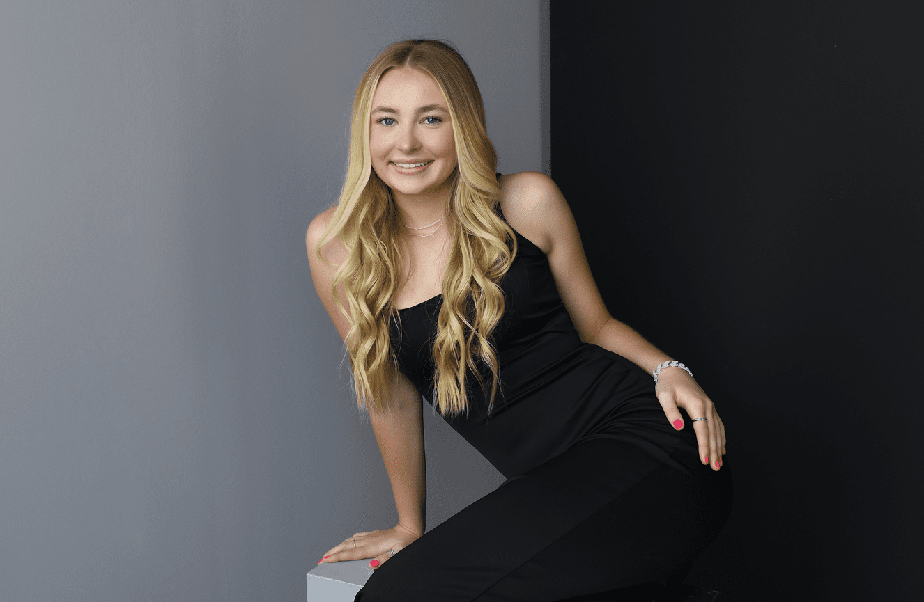 A young blonde woman in a black strap dress smiles at the camera.