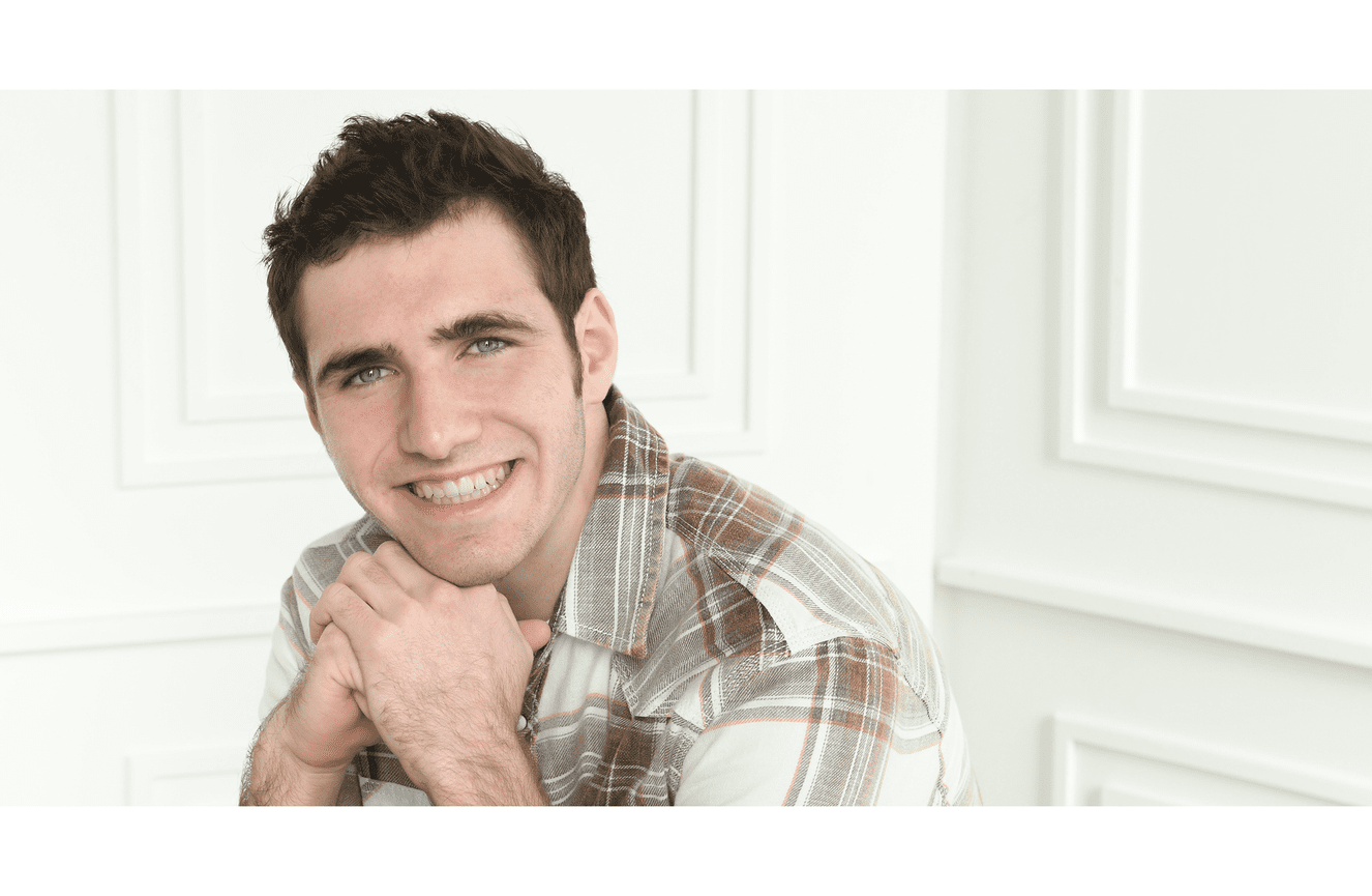 A senior boy in a plaid shirt smiling while resting his chin in his hands.