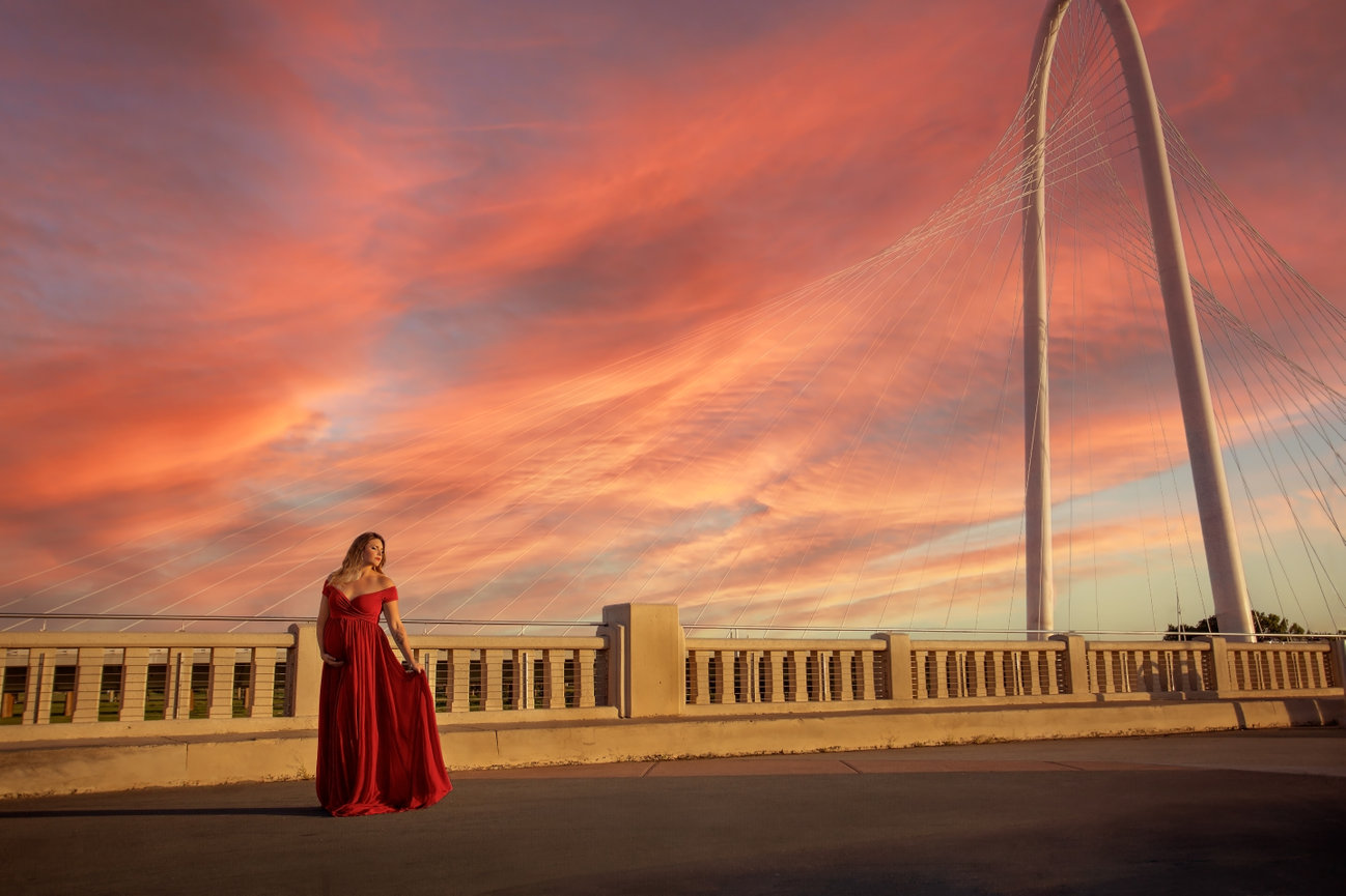 Dramatic maternity photo captured on a bridge in Dallas with iconic arched support in the background under a colorful afternoon sky.  Woman is wearing an elegant red gown.