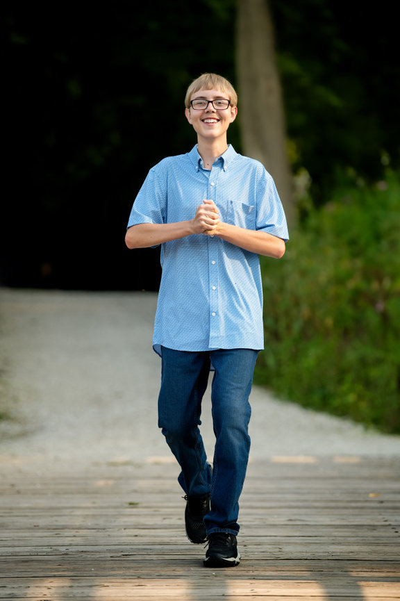 A westosha central high school senior boy walking on a wooden path, wearing glasses, a blue shirt, and jeans. Trees in the background at petrifying springs park.